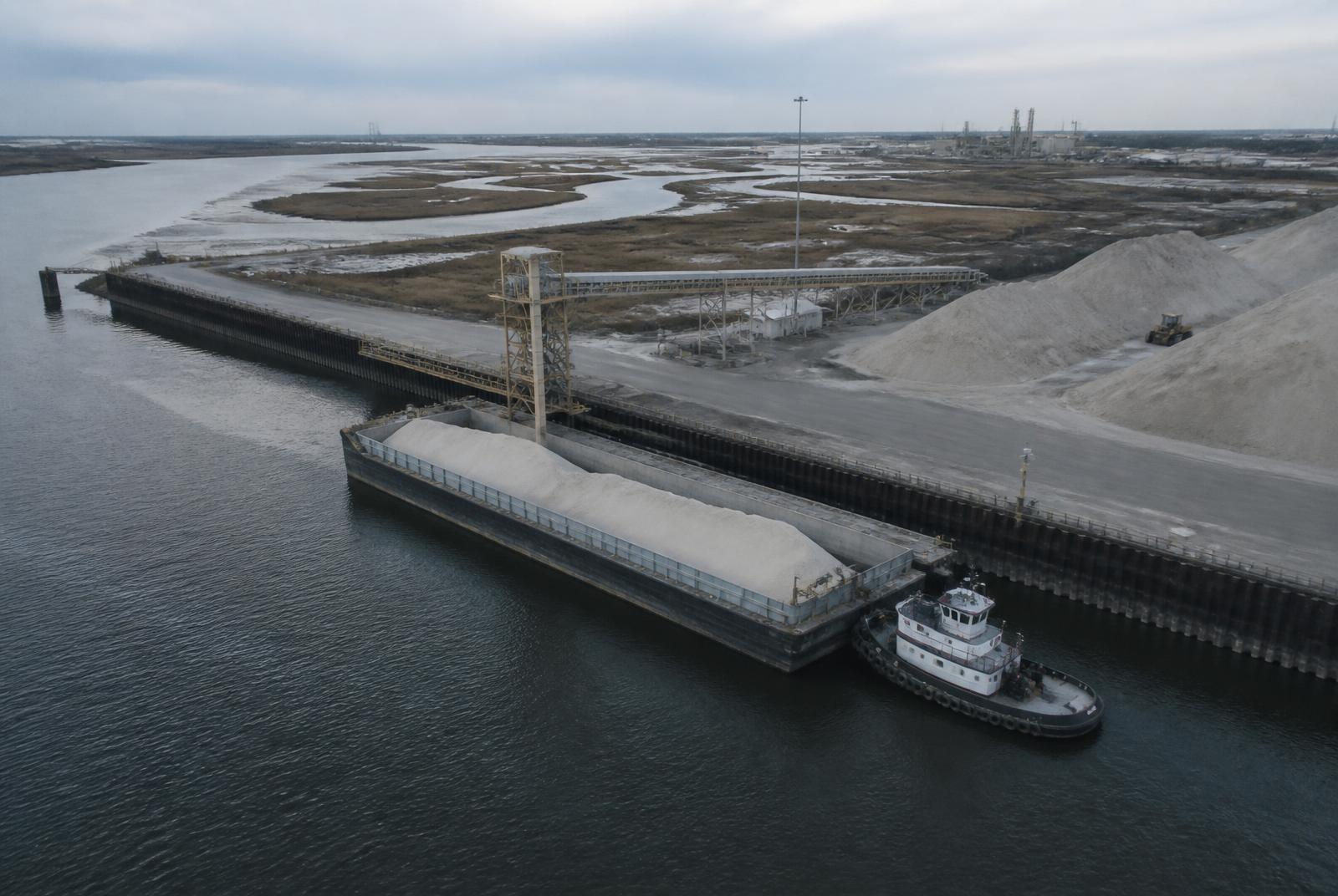 Aerial view of a working waterfront with a loaded barge, tug and bulk material handling conveyor