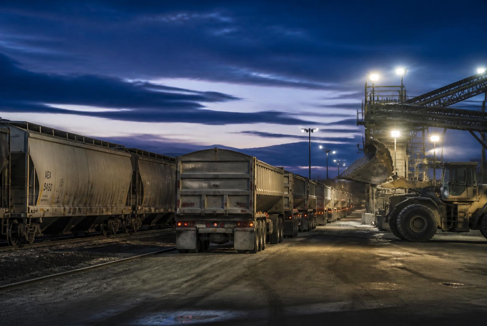 Line of bulk haul trailers and rail hopper cars at an industrial transload yard at dusk